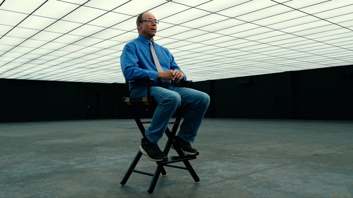 Steven Greer sitting on a folding chair in a large room with a ceiling tiled in panel lights