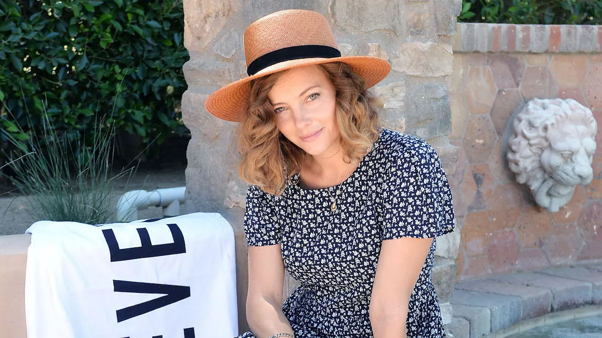 Bijou Phillips wearing a star hat and summer dress while sitting by the lip of a pool and looking at the camera