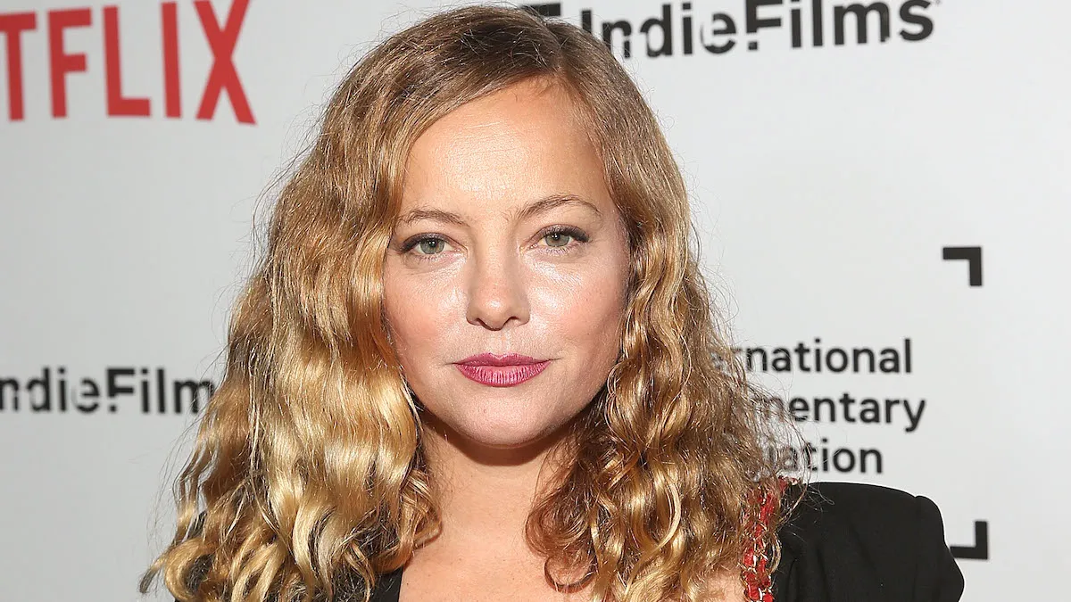 Bijou Phillips on the red carpet staring directly into the camera with curly hair, a red dress, and a black jacket