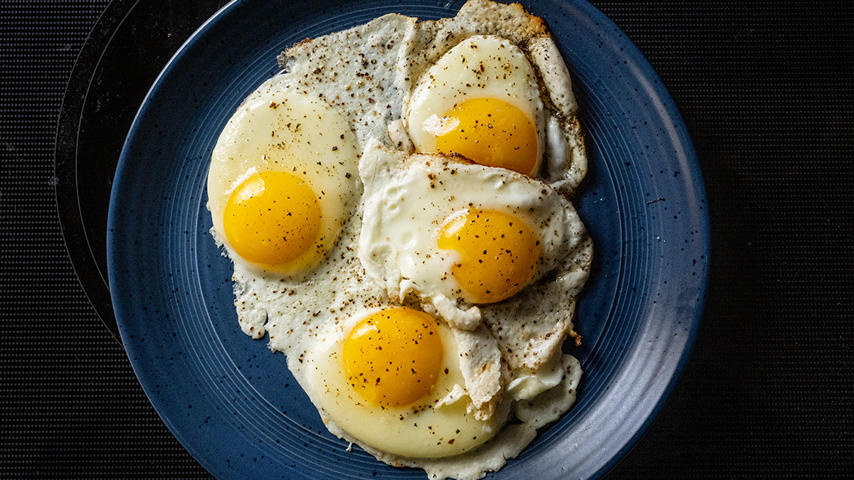 Four fried eggs cooked sunny side-up style and with runny style yokes with black pepper sit on a large blue plate.