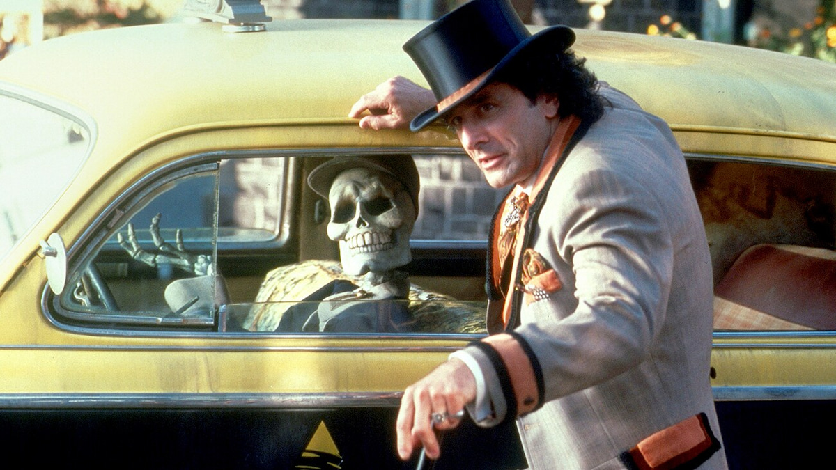 Kalabar from Halloweentown leans on a car driven by a skeleton. 