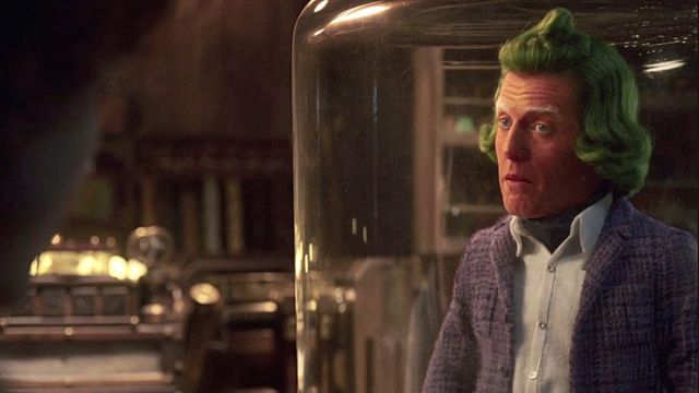 Hugh Grant in full costume, orange skin, green hair, and purple blazer, as an Oompa Loompa in the film 'Wonka'.