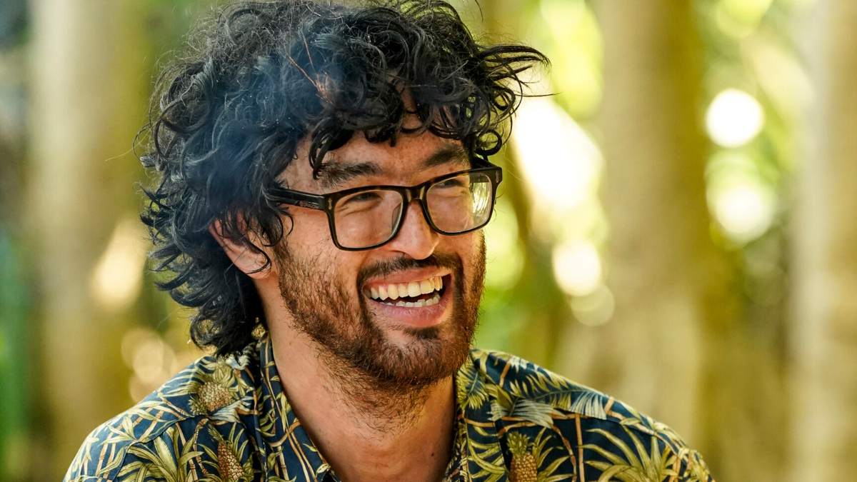 Matt Blankship is seen smiling at camp, wearing black glasses and a pineapple print button down shirt. 