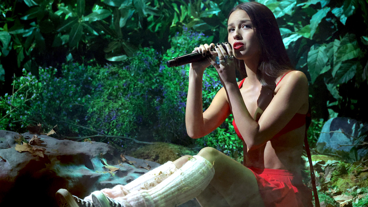 Olivia Rodrigo sitting on a floor covered in moss for her MTV performance, wearing a red top and skirt