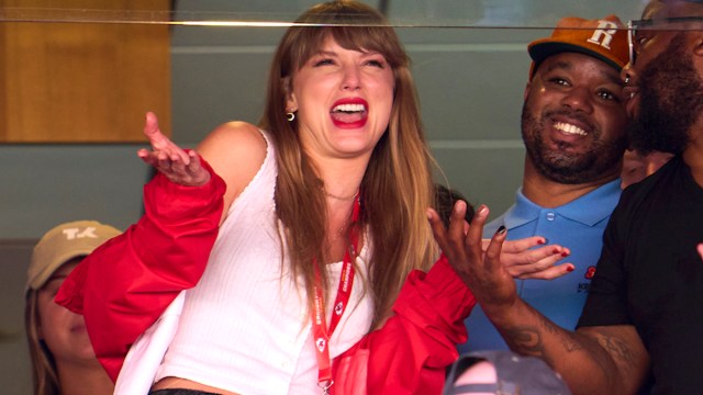 Taylor Swift shrugging and laughing while wearing a white shirt and red jacket in the box seats at the Kansas City Chiefs game