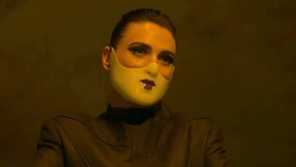 Katie McGrath como The Adjudicator, usando uma máscara na metade inferior do rosto