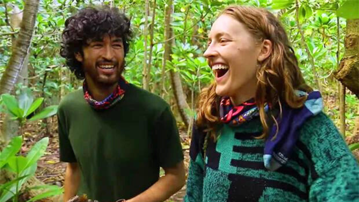 Matt Blankship and Frannie Marin are seen laughing with one another at camp. Both have buffs around their necks, while Matt is wearing a green tee shirt and Frannie is wearing a green poncho.