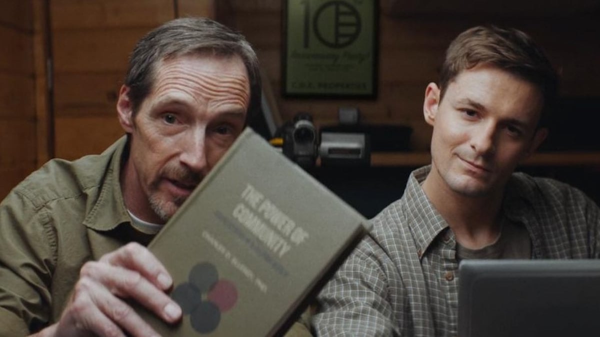Two men look at someone off-screen, one holding a book. 