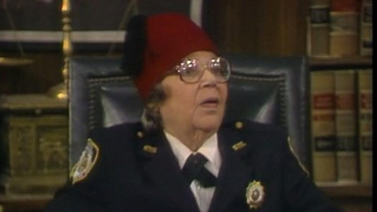 A woman in a police officer's outfit sits in a fancy leather chair in front of a bookcase full of old books.