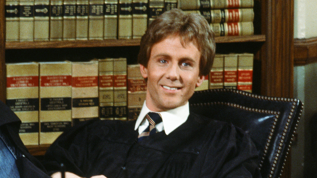 A man in judge's robes sits on a fancy leather chair in front of a bookshelf filled with law books.