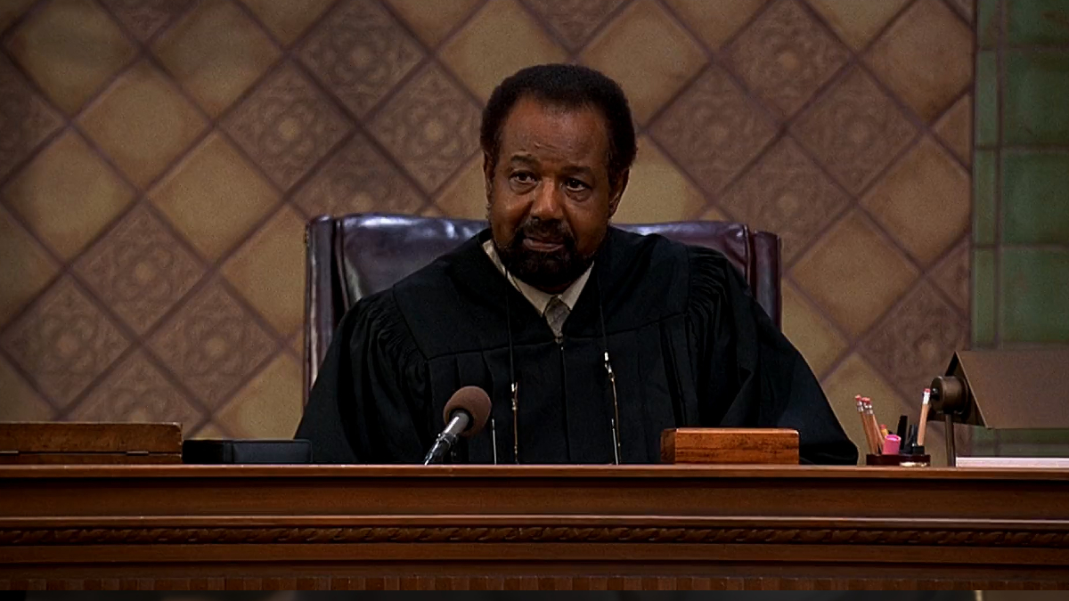 A black man in lawyers robes sits in a fancy leather chair while trying a case.
