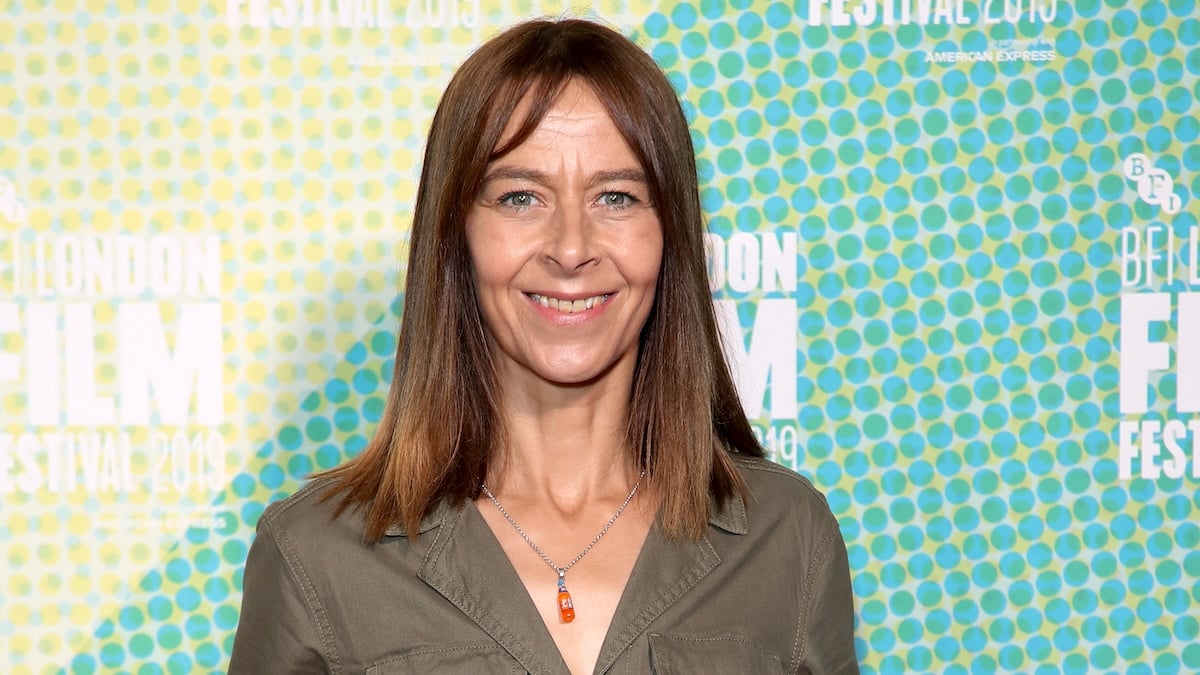 Kate Dickie standing in front of a green and yellow background wearing a forest green button up jacket