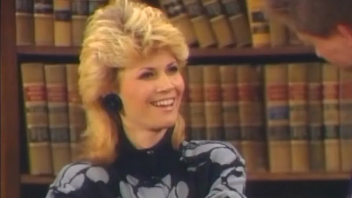 A blond woman sits in front of a large bookshelf filled with law books.