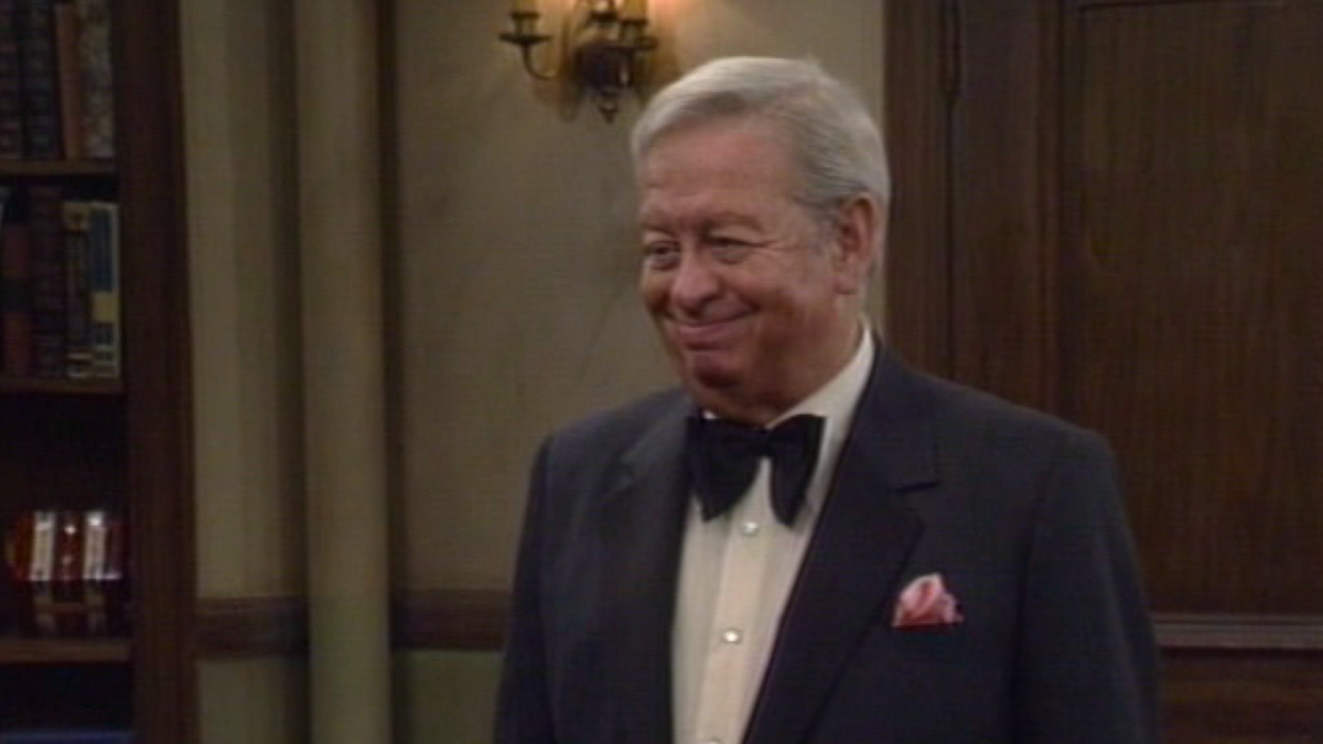 A old man stands in a tuxedo suit and bow tie.
