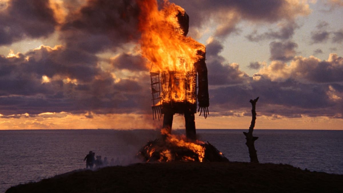 A group of people stand on the shore, watching a large burning wooden/wicker man.