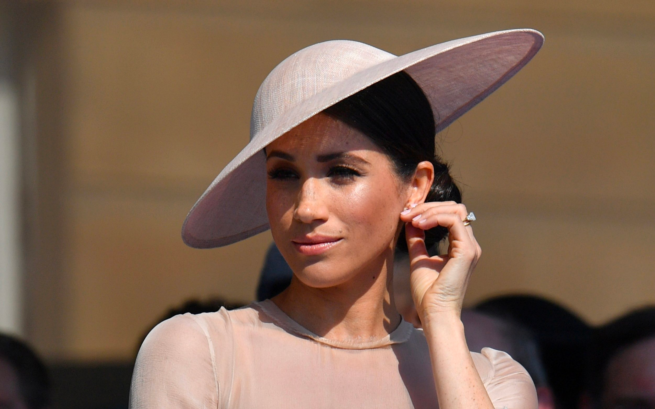 Meghan Markle is wearing a hat and holding her ear. 