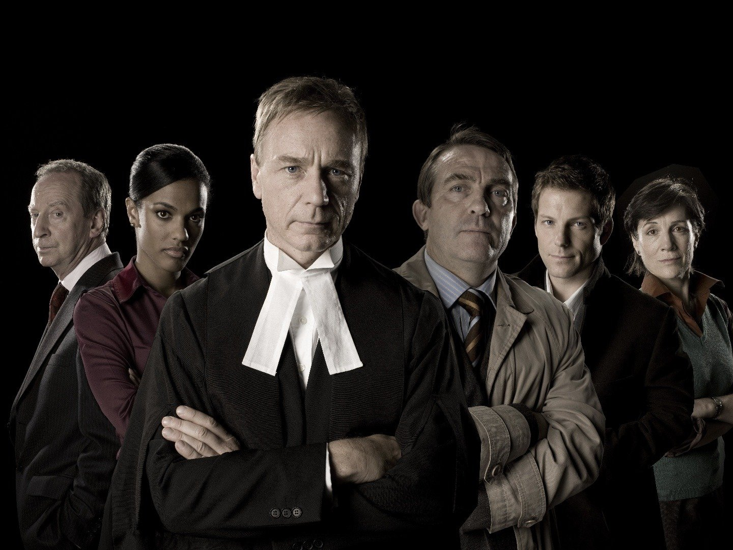 The cast of Law & Order: UK stands with their arms crossed in front of a black background.