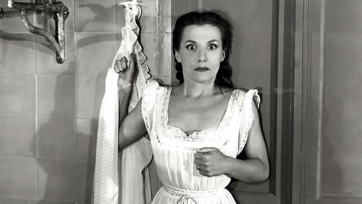 Woman in Les Diaboliques film stands in front of shower, gripping curtain in fear.