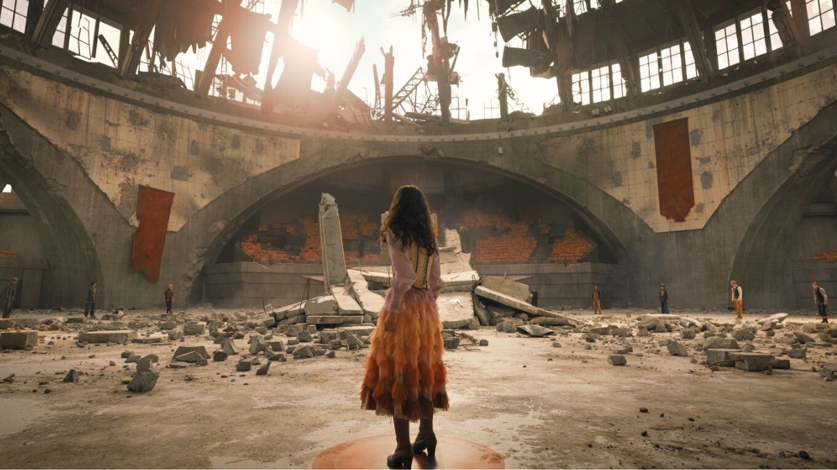 Rachel Zegler, who portrays Lucy Gray Baird, stands in the center of the 10th Hunger Games arena, her back turned toward us. In the background, many of the tributes are visible.