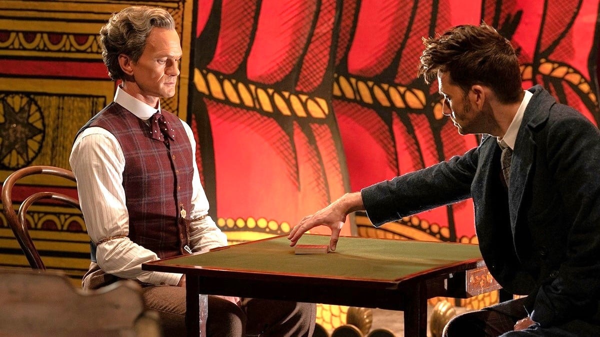 Neil Patrick Harris' Toymaker and David Tennant's Doctor have a tense face-off across a table in front of a stage in Doctor Who: The Giggle. 