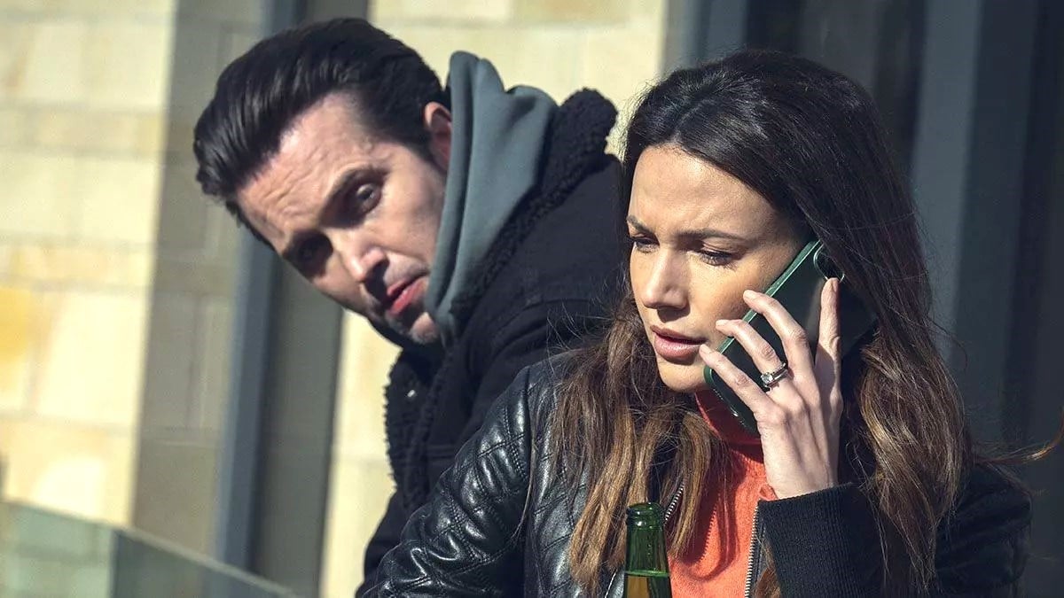Maya (Michelle Keegan) answers the phone as Shane (Emmett J. Scanlan) stands over her shoulder in Netflix's Fool Me Once.