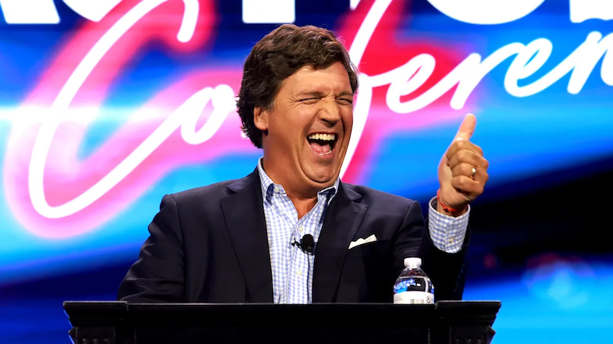 Tucker Carlson laughing and giving a thumbs-up at the Turning Point Action conference