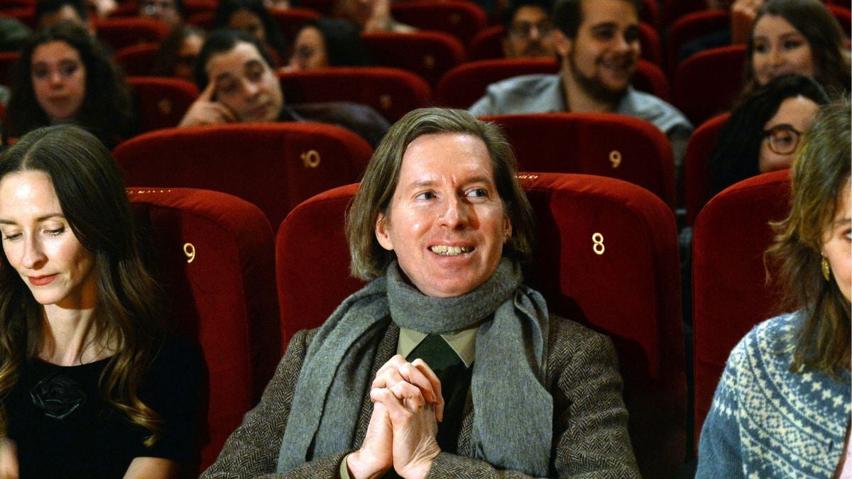 Wes Anderson sits in a movie theater, hands folded tag, wide grin on his face, amongst an excited crowd of moviegoers.