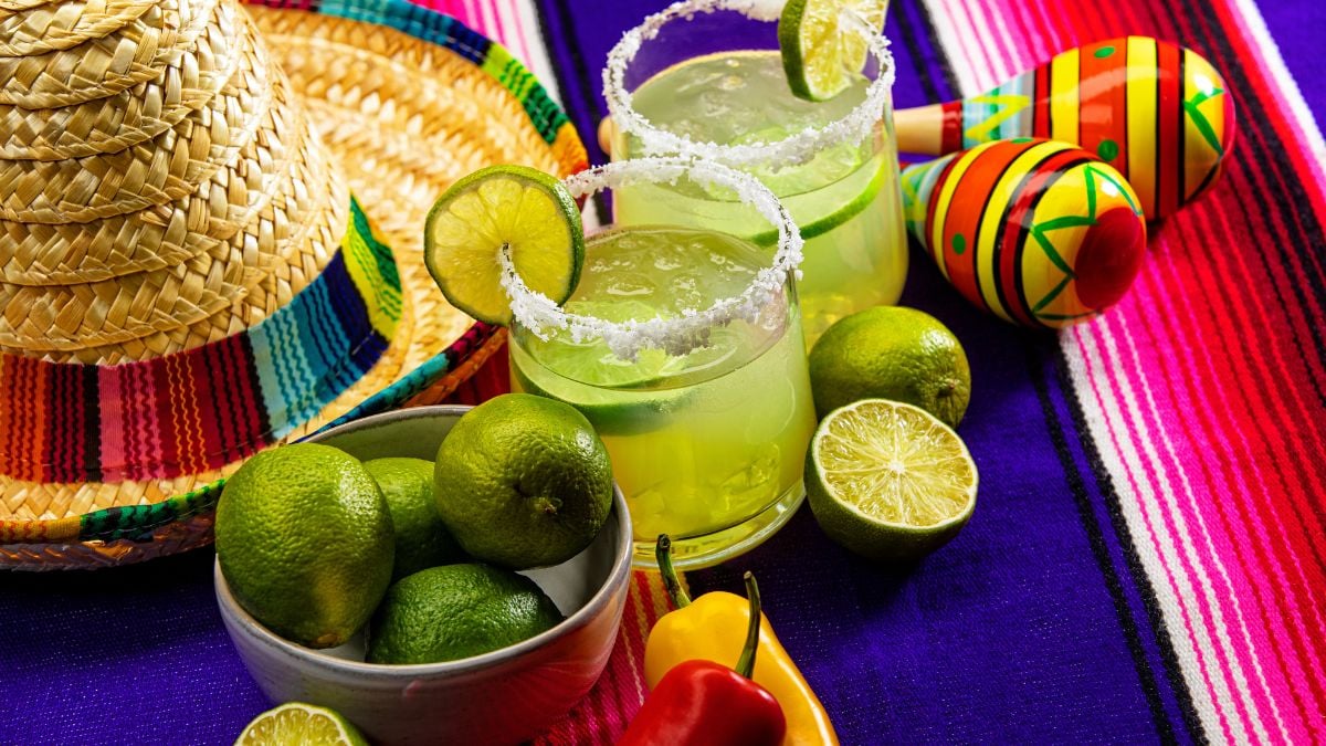 This is a photograph of two modern margarita glass with a rim of salt surrounded by fresh cut limes and chilis on a colorful striped mexican blanket next to a sombrero