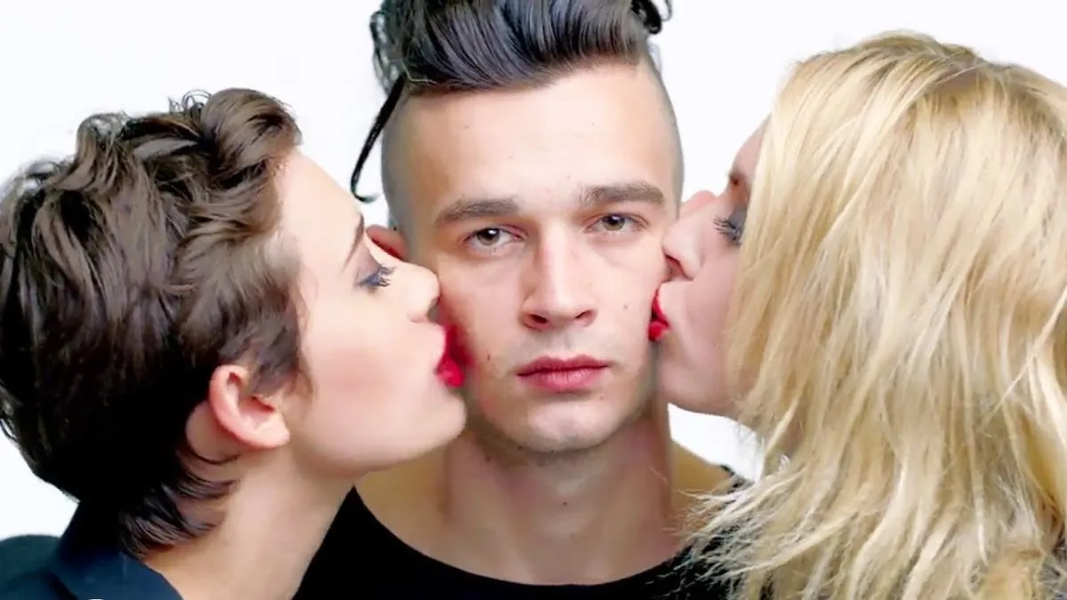 Matty Healy in The 1975's music video "Girls" with two women kissing his cheeks