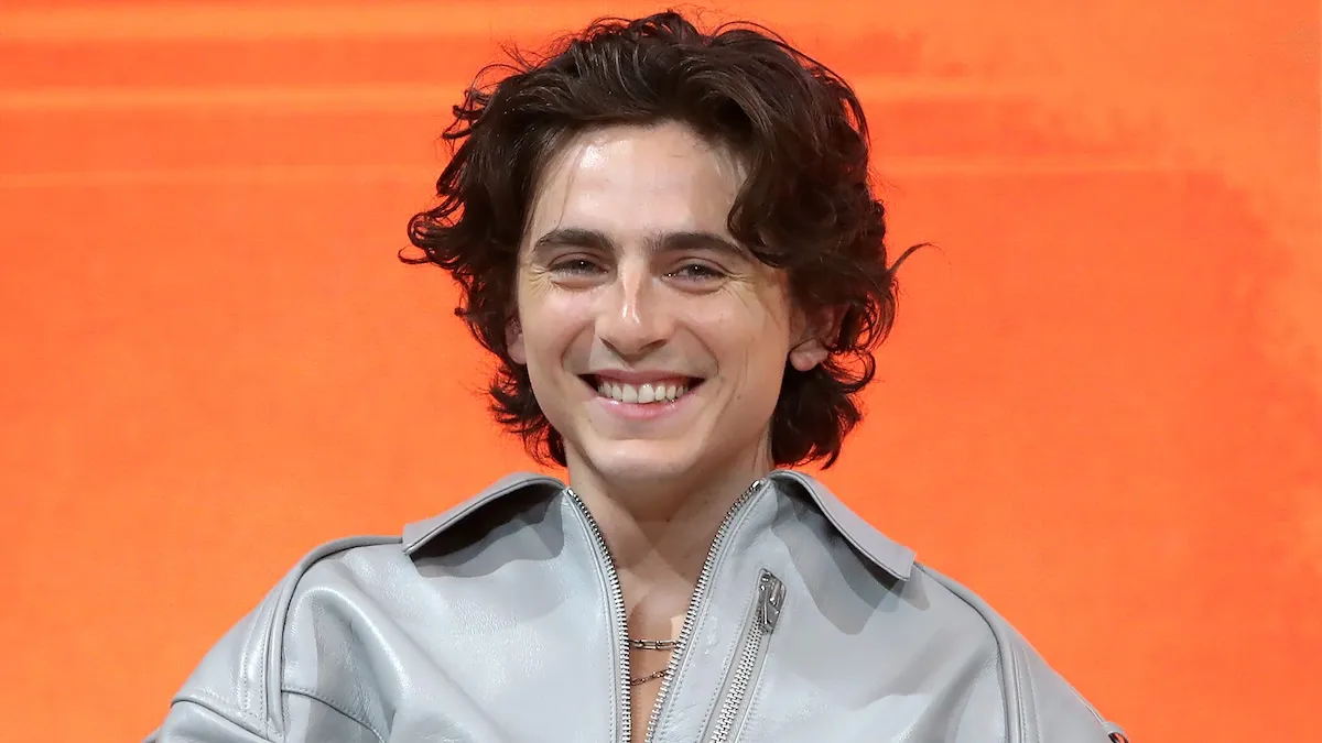 Timothee Chalamet smiling big in a silver jacket, backdropped by an orange backdrop.