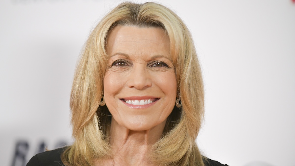 Vanna White on the red carpet, backdropped by a cream-colored backdrop. She is smiling at the camera