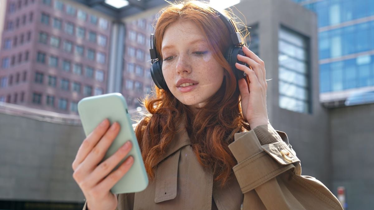 Teen redhead girl standing on urban street skyscrapers background wearing headphones using smartphone listening to music, audio book, travel guide or podcast while walking in big city.