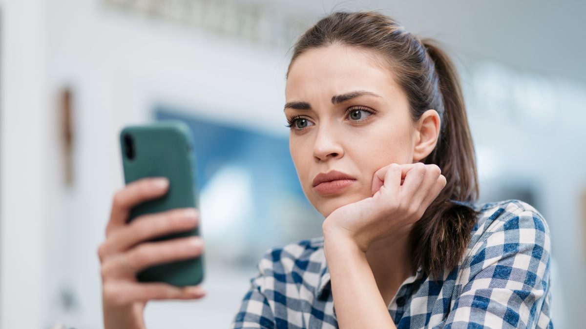 Close up of a depressed Caucasian young woman sitting at home, reading some bad news on her smart phone, feeling sad and worried