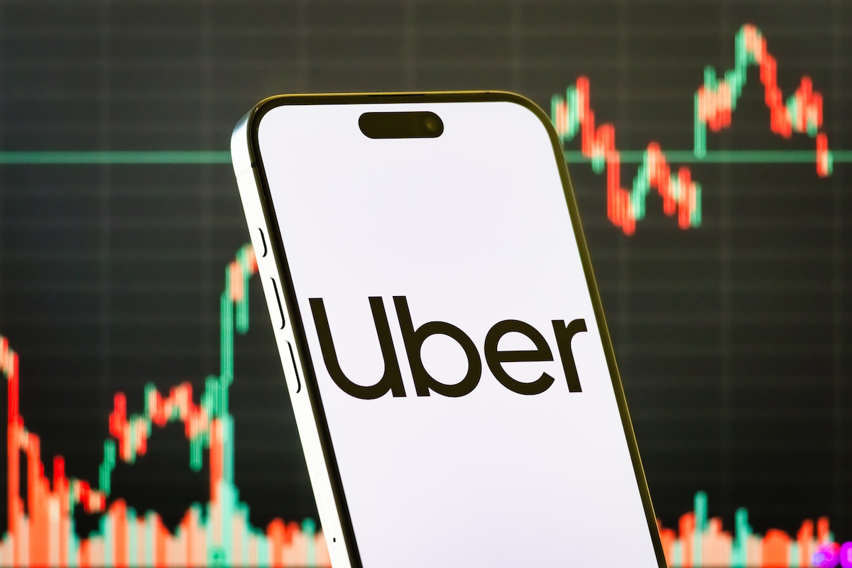 CHONGQING, CHINA - JULY 31: In this photo illustration, a smartphone displays the logo of Uber Technologies Inc. (NYSE: UBER), a global ride-hailing and mobility-as-a-service company, in front of a screen showing the company’s latest stock market chart on July 31, 2025 in Chongqing, China.