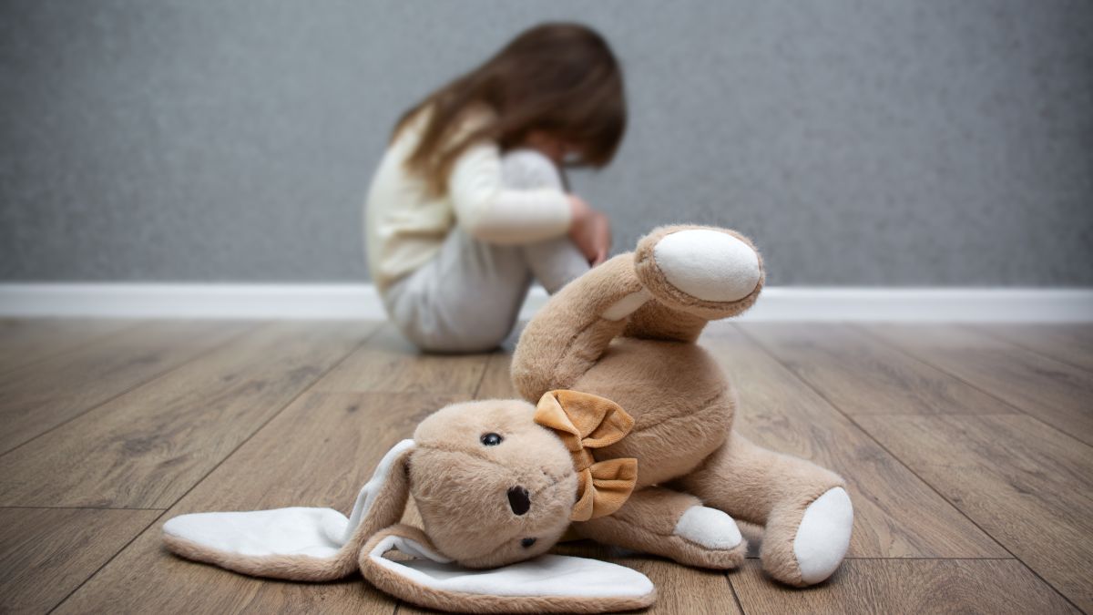 A stuffed bunny on the floor, a scared child sitting behind it. Child abuse. Bullying in the family.