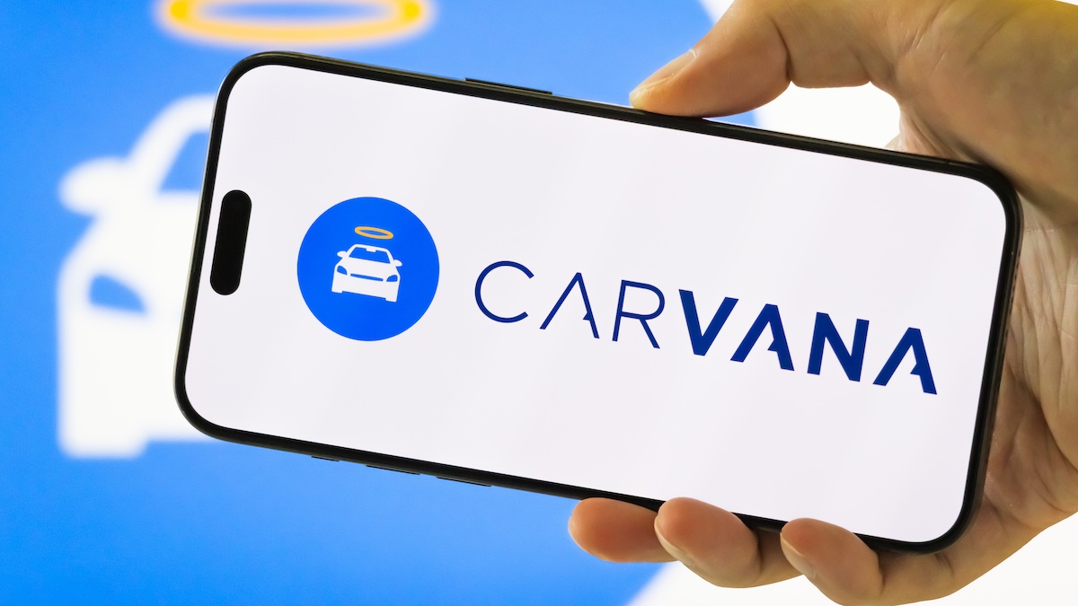 CHONGQING, CHINA - JULY 28: In this photo illustration, a person holds a smartphone displaying the logo of Carvana Co. (NYSE: CVNA), an American e-commerce platform for buying and selling used cars, with the company’s signature halo car emblem visible in the background, on July 28, 2025 in Chongqing, China.