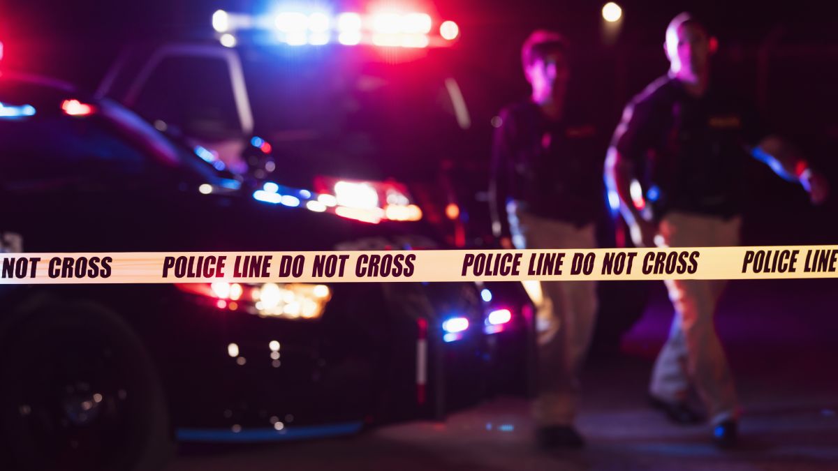 wo multi-ethnic police officers standing in front of patrol cars, behind police tape, wearing bulletproof vests and duty belts. The policewoman is a mature African-American woman in her 40s. Her partner is a mid adult man in his 30s. It is nighttime and the emergency lights on top of the vehicles are flashing. The focus is on the cordon tape in the foreground.