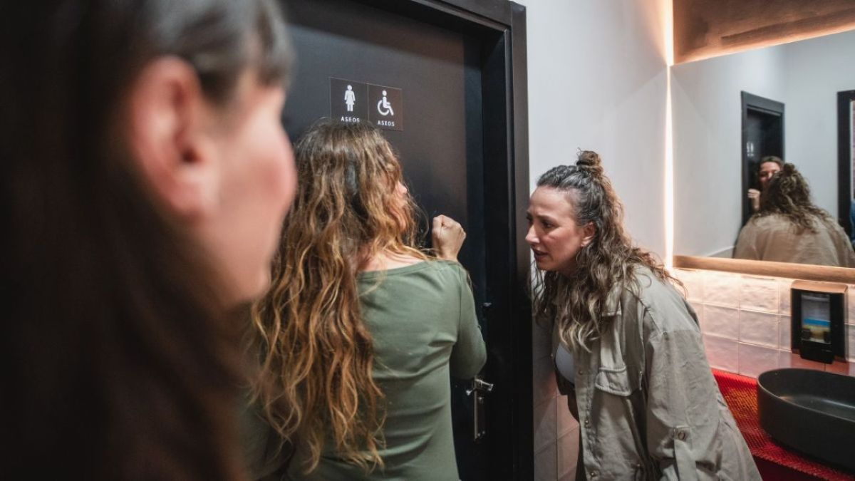 Women queueing outside bathroom door and knocking, showing urgency